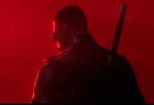 A darkly dressed man with a sword on his back, seen from behind against red light.