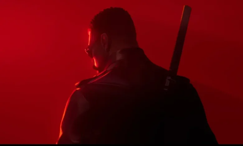 A darkly dressed man with a sword on his back, seen from behind against red light.