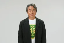 Shigeru Miyamoto stands against a plain background wearing a white shirt with a green Zelda anniversary design.