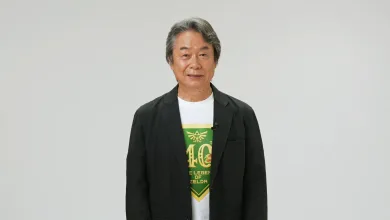 Shigeru Miyamoto stands against a plain background wearing a white shirt with a green Zelda anniversary design.