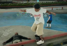 A skater in a white shirt and tan pants balances on a skateboard while grinding a low rail at a skatepark, with another character on the ground nearby.