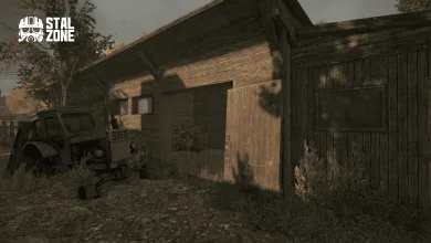 A weathered wooden barn with an open door stands beside an old vehicle in overgrown surroundings.