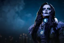 A dark-haired contestant with skull-like makeup and a glowing blue pendant stands against a smoky blue background.