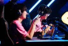 Two esports players in pink jerseys sit at gaming computers under blue stage lighting.