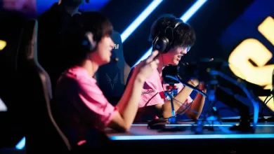 Two esports players in pink jerseys sit at gaming computers under blue stage lighting.
