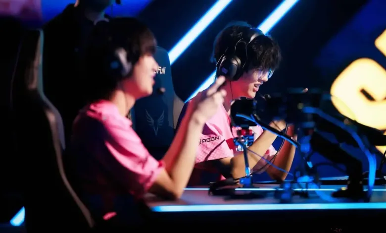 Two esports players in pink jerseys sit at gaming computers under blue stage lighting.