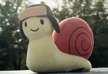 The Gaijin Snail plush sits on a ledge outdoors, with trees blurred in the background.