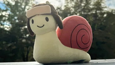 The Gaijin Snail plush sits on a ledge outdoors, with trees blurred in the background.