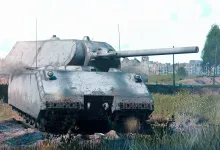 A large gray Maus tank sits in a grassy field with buildings in the background.