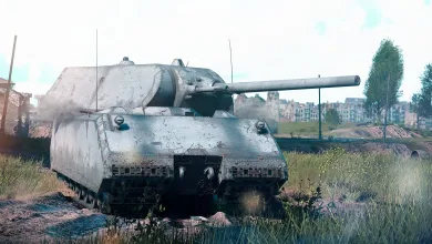 A large gray Maus tank sits in a grassy field with buildings in the background.