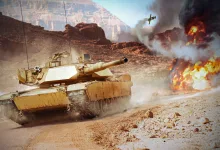 A tan tank drives across rocky desert terrain as smoke and flames rise nearby.